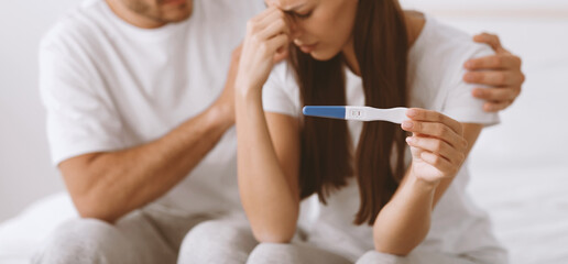 Cropped disappointed sad young caucasian man support his wife, hold pregnancy test, sit on bed in bedroom interior, panorama. Unwanted pregnancy, infertility, health problems and crisis in family