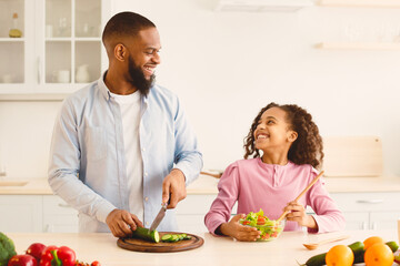 Portrait Of Smiling Black Father And Daughter Cooking Healthy Vegetarian Food In Kitchen, Afro Family Preparing Vegetable Salad For Lunch At Home, Enjoying Homemade Food, Looking At Each Other