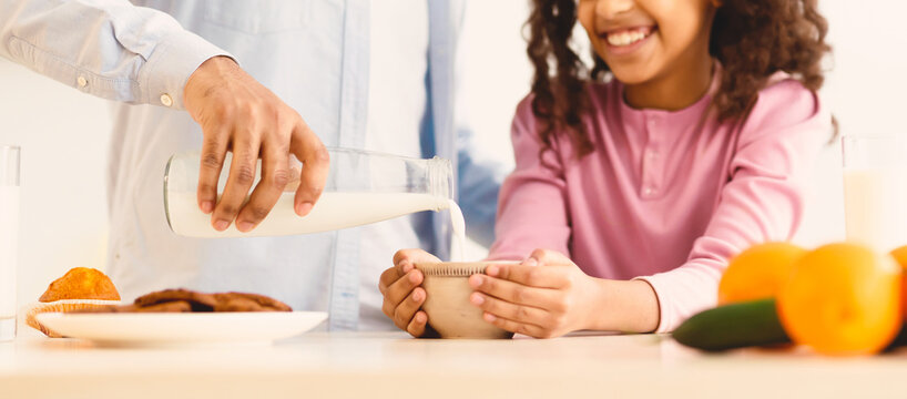 Nutrition And Breakfast. Closeup of black man pouring fresh milk from bottle into daughter's bowl with cereals. Happy curly girl having delicious meal together with daddy in kitchen, selective focus - Powered by Adobe