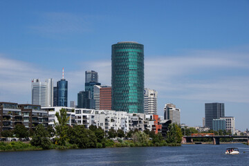Fototapeta premium Modern city skyline featuring a unique green glass skyscraper, residential buildings, and a serene river, showcasing urban architecture and vibrant city life