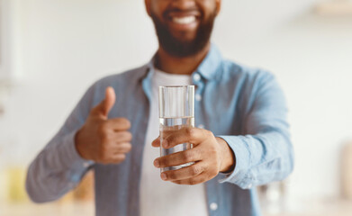 Smiling young black man with beard enjoy good morning, show glass of clean water and thumb up on minimalist kitchen interior, close up, blurred. Good habit, healthy lifestyle, hydration and drink aqua