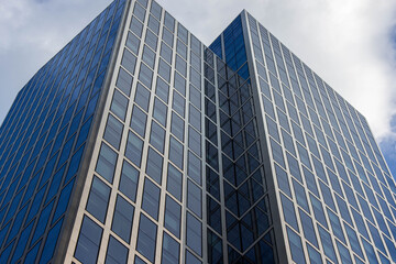 Obraz premium Modern glass skyscraper with reflective windows and geometric design, showcasing urban architecture against a cloudy sky, representing contemporary city life and innovation