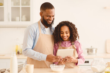 Cooking Together. Portrait of happy black father and daughter in aprons cooking in the kitchen, holding and using digital tablet, watching food video blog or reading recipe online on culinary website