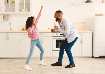 Family Holiday And Togetherness. Joyful black man and his excited daughter dancing and having fun together at the kitchen, smiling dad singing song in whisk as a mic, celebrating father's day
