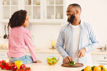 Happy Loving Family. Cute Curly Black Girl Sitting On Dinner Table, Father Slicing Cucumber On Cutting Board With Knife. Man Spending Time With Daughter In The Kitchen, Talking And Cooking Together