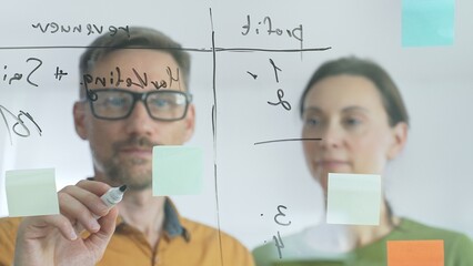 Business team of professionals brainstorming near glass board, using sticky notes for strategic planning, writing calculations, sharing innovative ideas during collaborative work