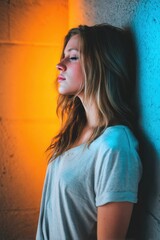 Fototapeta premium A young woman with flowing hair leans against a textured wall, illuminated by warm orange and cool blue lights, enhancing the mood and visual appeal of the scene