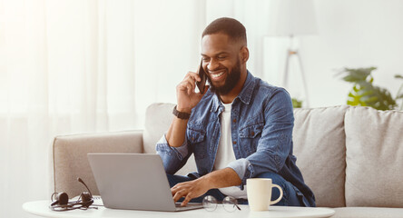 Naklejka premium Happy millennial african american male work on laptop and talk by phone at workplace on couch in living room interior. Success home business, distant communication and new normal due covid-19 pandemic