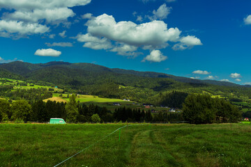 Bright rural panorama with scattered farmhouses, forested hills, and open pastures under a blue sky with soft clouds. A peaceful countryside view showing the harmony between nature and rural life.
