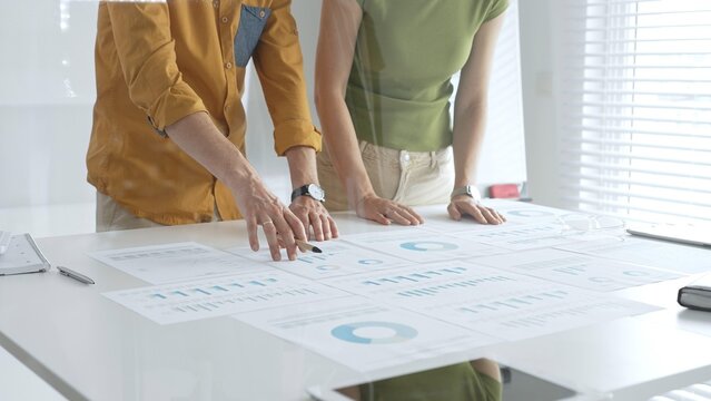 Two business professionals are examining financial data and reports, pointing at charts and graphs displayed on a table during a collaborative office meeting