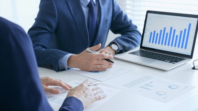 Business colleagues discussing financial data on a laptop and printed documents while analyzing charts and market trends. Business people and finance concept