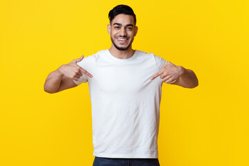 Cheerful arab guy pointing with both hands at his blank white t-shirt and smiling, posing over...