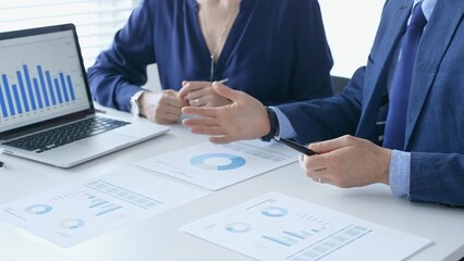 Businessman and businesswoman are analyzing financial data on a laptop and printed documents, pointing at charts and graphs during a meeting in bright office. Business people and finance concept