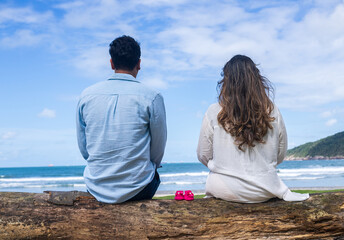 Fototapeta premium couple looking at the sea sitting on a tree trunk with baby shoes, first-time mother and father