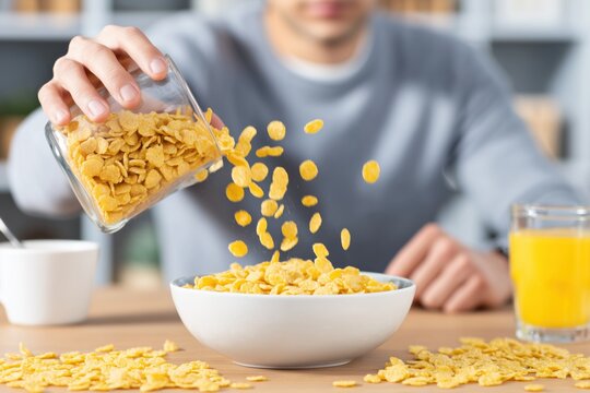 Man pouring cornflakes into a white bowl at breakfast with a glass of orange juice nearby