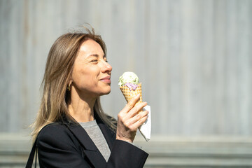 Happy businesswoman eating ice cream walking outside during lunch break on sunny day. Portrait of content female strolling with on the go dessert, time with yourself, indulgence with unhealthy snack