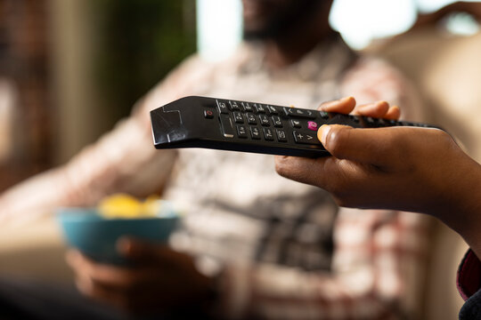 Closeup of female hand with thumb on black remote pointed at television, adjusting volume. African American couple sitting together watching tv shows or movies, relaxing in modern living room. - Powered by Adobe
