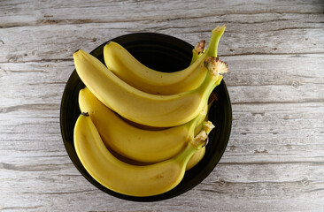 A collection of bright yellow bananas rests in a sleek black bowl atop a rustic wooden table. The lighting accentuates their vibrant color, inviting a closer look