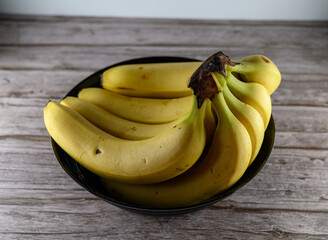 A collection of bright yellow bananas rests in a sleek black bowl atop a rustic wooden table. The lighting accentuates their vibrant color, inviting a closer look