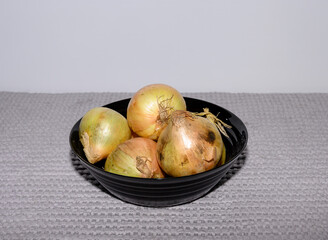 A black bowl filled with fresh, yellow onions rests on a gray textured surface. The lighting highlights the onions' natural colors and textures, creating a vibrant look