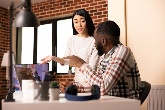 Asian woman and black man review coding work on laptop together in apartment. Young diverse couple work from home, collaborating on web app development, creating prototype for startup tech company. - Powered by Adobe