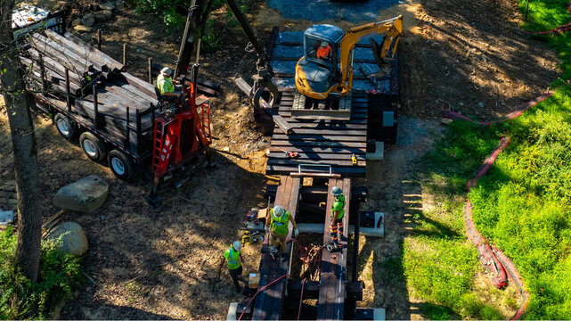 A construction crew collaborates to install a timber bridge in a forested region. Heavy equipment and logs are positioned as workers coordinate their efforts under clear blue skies.