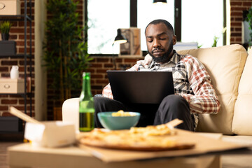 African american man seated on couch with serious expression, working on laptop during downtime. Male entrepreneur surrounded by snacks and drink, completes company tasks in brick wall home.