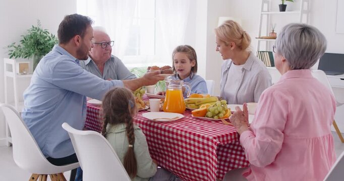 Multi generational family sits at table enjoying bright atmosphere of breakfast. Grandparents, young husband and wife and their children eat fruit together and drink fresh orange juice in bright room.