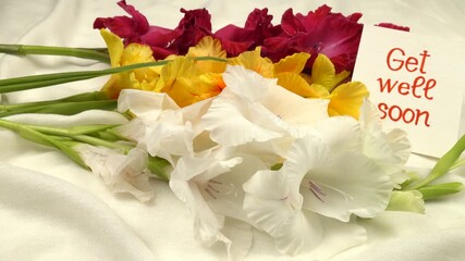 A bouquet of multi-colored gladioli and a card with a wish for a speedy recovery «Get well soon" on white fabric