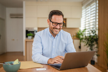 Obraz premium Smiling businessman working from home using laptop at the table
