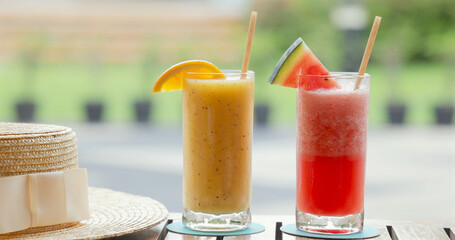 Summer patio scene with refreshing tropical smoothies. Orange and watermelon flavors in tall glasses beside straw hat. Perfect outdoor relaxation concept.
