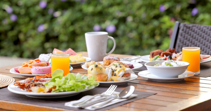 Outdoor brunch table with fresh breakfast dishes, juice, coffee set on wooden table in garden setting. Relaxation, weekend leisure, al fresco dining concept. Vacation, hotel.
