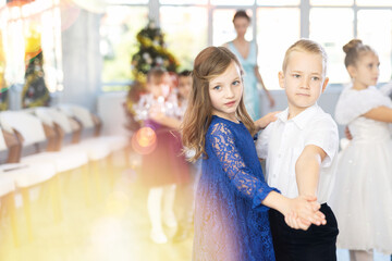 Couple of smiling tweens, boy and girl in festive clothing performing graceful waltz with group classmates under guidance of female teacher during Christmas event at school..