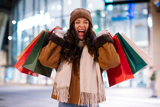 Overjoyed European lady holding shopper bags and screaming with happiness after successful shopping posing outdoors in front of mall in the evening