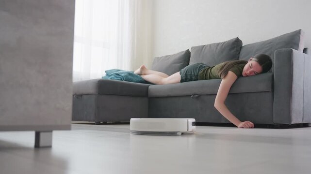 Woman lies on gray couch with relaxed posture, hand resting on floor as robot vacuum cleaner passes by, illustrating smart home cleaning, effortless automation, and modern lifestyle comfort indoors