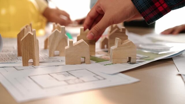 Architect and engineer discuss about building blueprint on table with architectural document, measurement tools scatter around at office, use wooden block for house positioning. Closeup. Delineation.