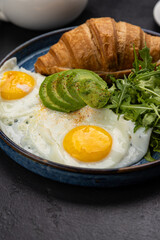 Healthy breakfast with croissant, avocado, sunny side eggs and salad on dark background