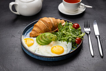 Healthy breakfast with croissant, avocado, sunny side eggs and salad on dark background
