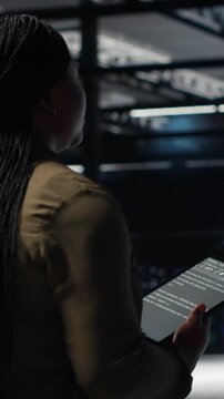 Vertical video Engineer in data center using tablet to deploy secure server architecture. African american woman in server hub using device to do rollout of cybersecurity framework, camera B