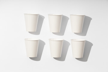 Pattern of several white paper cups for drinks on white background with hard light. Ecological disposable tableware for coffee, tea or water. Taken from a top view and flat lay.