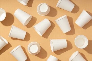 Pattern of several white paper cups for drinks on beige background with hard light. Ecological disposable tableware for coffee, tea or water. Taken from a top view and flat lay.