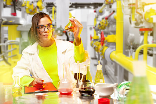 Female scientist in a lab coat analyzes a chemical solution in a flask, surrounded by laboratory glassware. Industrial background highlights connection between science and industry. - Powered by Adobe
