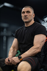athletic man sits on a bench in a gym taking a moment to rest. He appears focused and determined after completing a challenging workout. The environment suggests a dedicated training space.