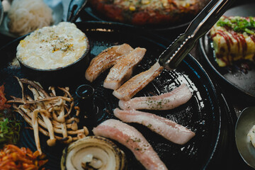 Sizzling Korean BBQ pork belly (samgyeopsal) on tabletop charcoal grill with rising smoke and traditional banchan side dishes, close-up shot in cozy restaurant setting with copy space.