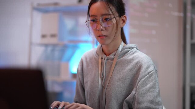 A female programmer debugs code on her laptop in a tech hub. In the background, a screen displays data streams, representing a complex software development or data science environment.