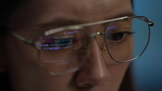 An extreme close-up of a young woman's glasses reflecting computer code. She is working late at night, focused on a complex technology problem on her screen.