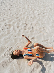 Sunlit beach scene featuring a person in a striped bikini lying on soft white sand, relaxed pose, warm light, and a carefree summer vibe.