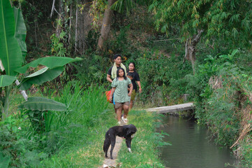 A father, mother, son, and daughter walk along a narrow path beside irrigation water, heading home through the green tropical countryside after bathing.