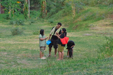 Fototapeta premium A cheerful family gathers in a green meadow where the mother shows wildflowers to the father while the boy happily plays with their dog.