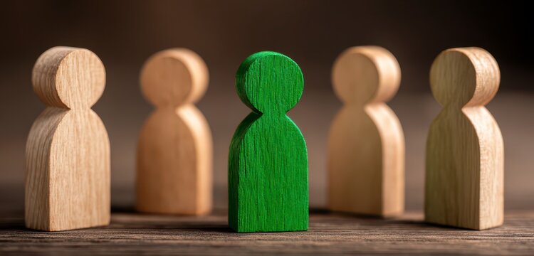 A single green wooden figurine stands out amongst a group of beige wooden figurines on a dark wooden surface